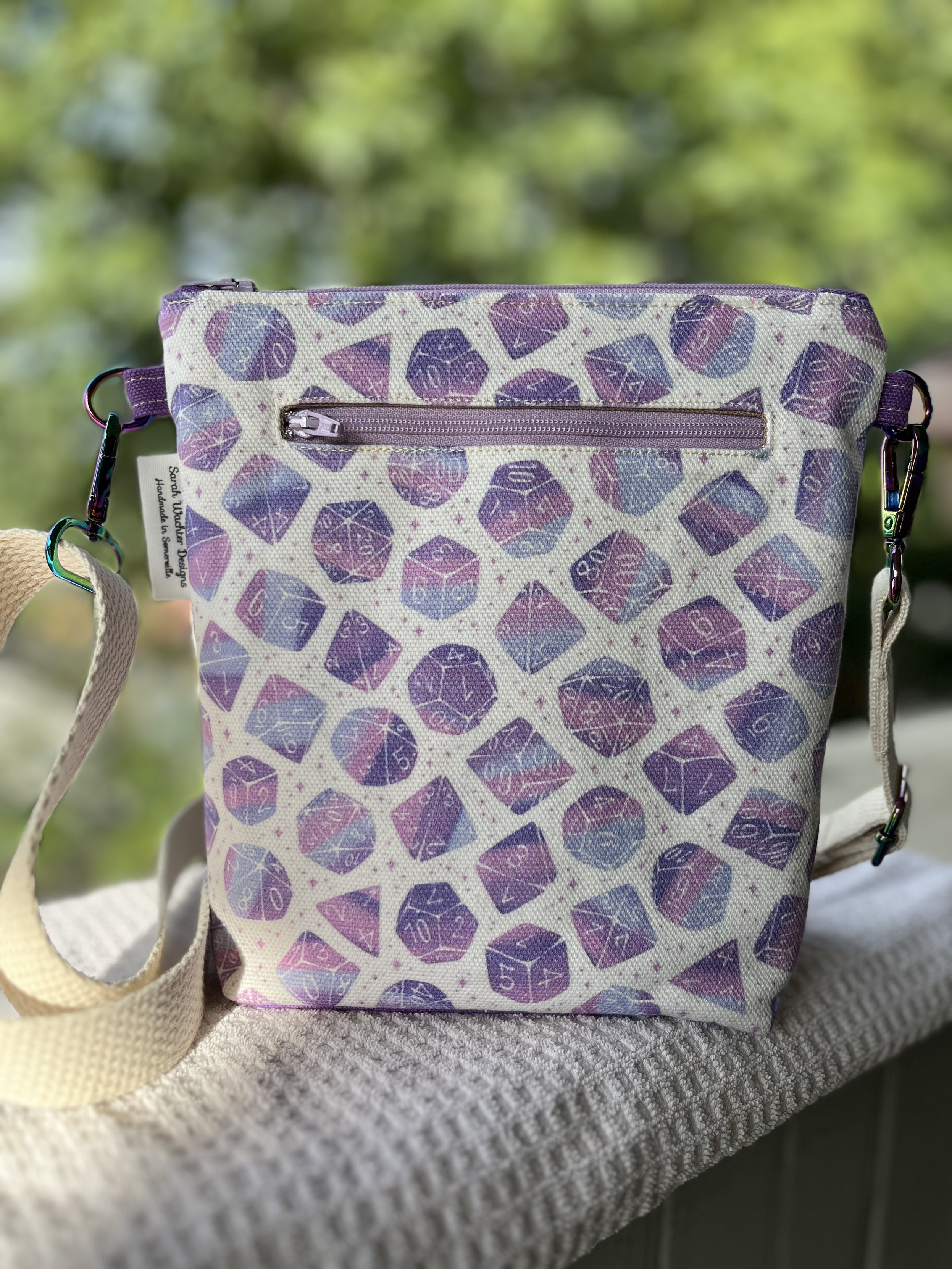 A crossbody bag with TTRPG dice scattered over it, in an ombre pink-blue-purple colorway.
