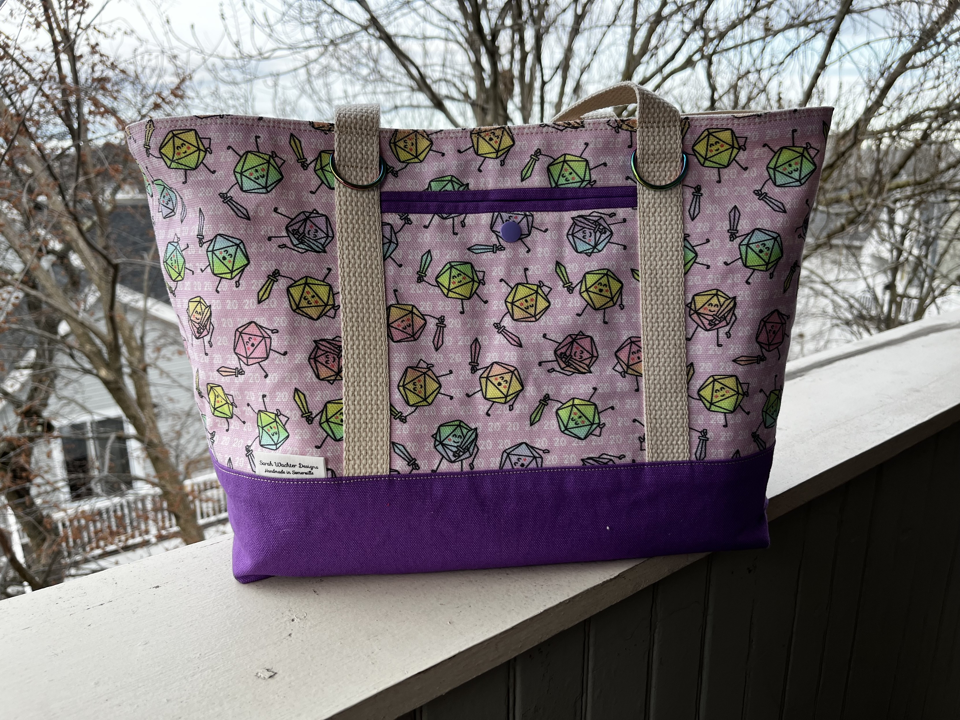 A tote bag with a pink background and lavender base, with a pattern of cute D20s holding swords scattered across it.  The D20s have a rainbow gradient thing going on.  It's super cute.