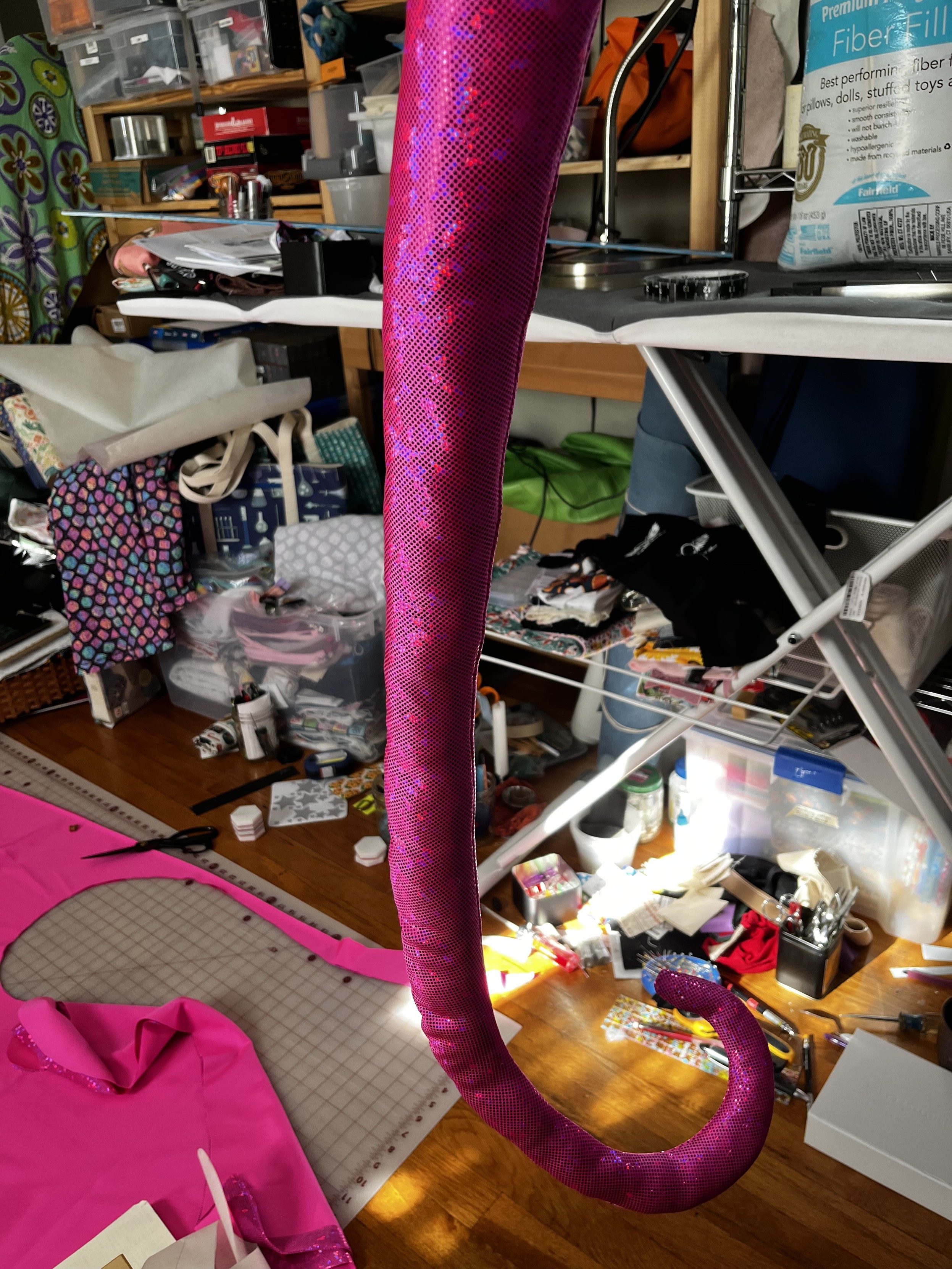 An aggressively pink tentacle made out of hot pink glitterball fabric hands in front of a very messy sewing room. The tentacle curls at the end and is delightful.