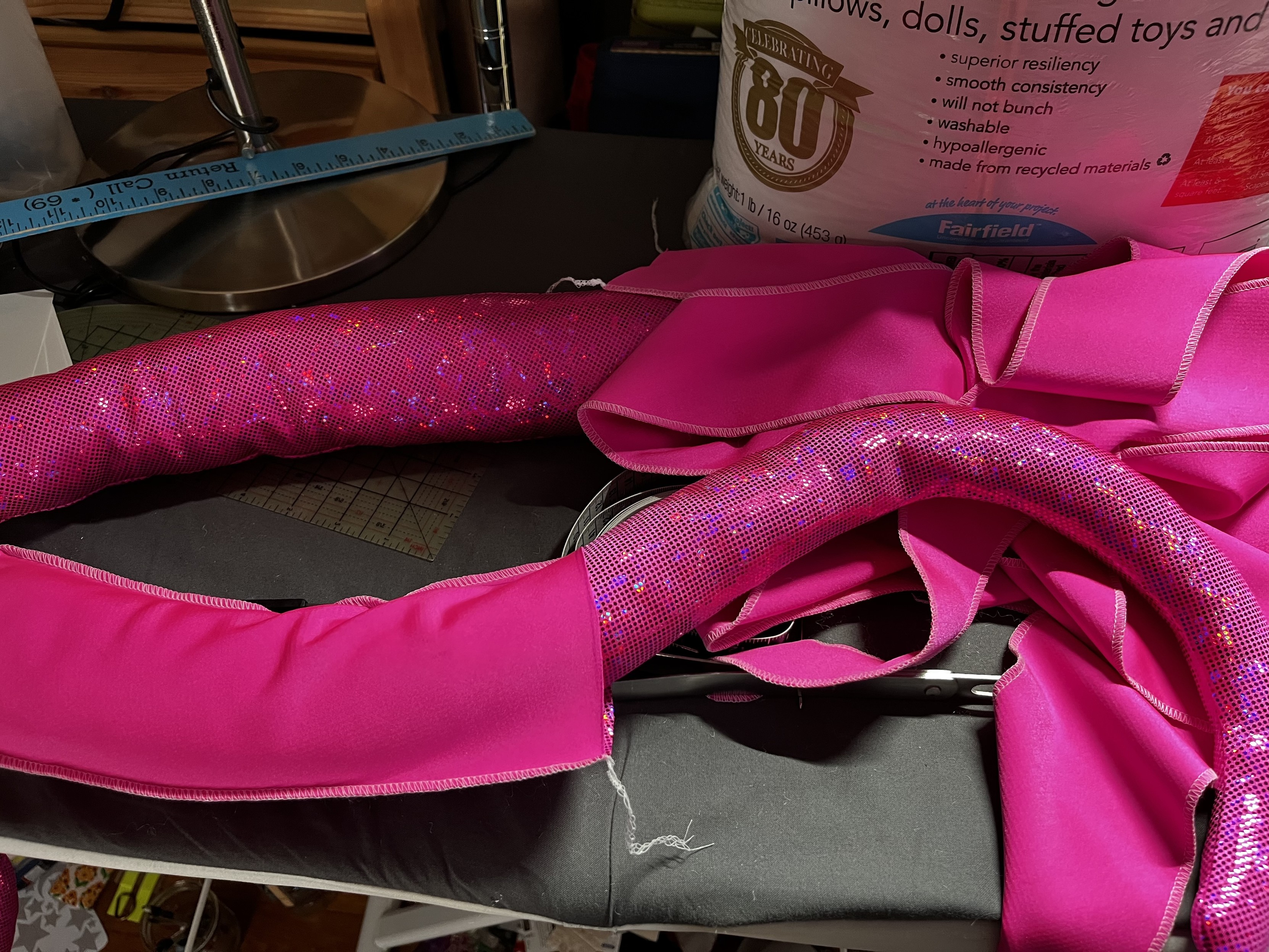 A hot pink sparkly tentacle on an ironing board with a bag of polyfil in the background. The tentacle is partially inverted, with the end of the tentacle stuffed and right side out, while the wider part is still inside out and folded over.
