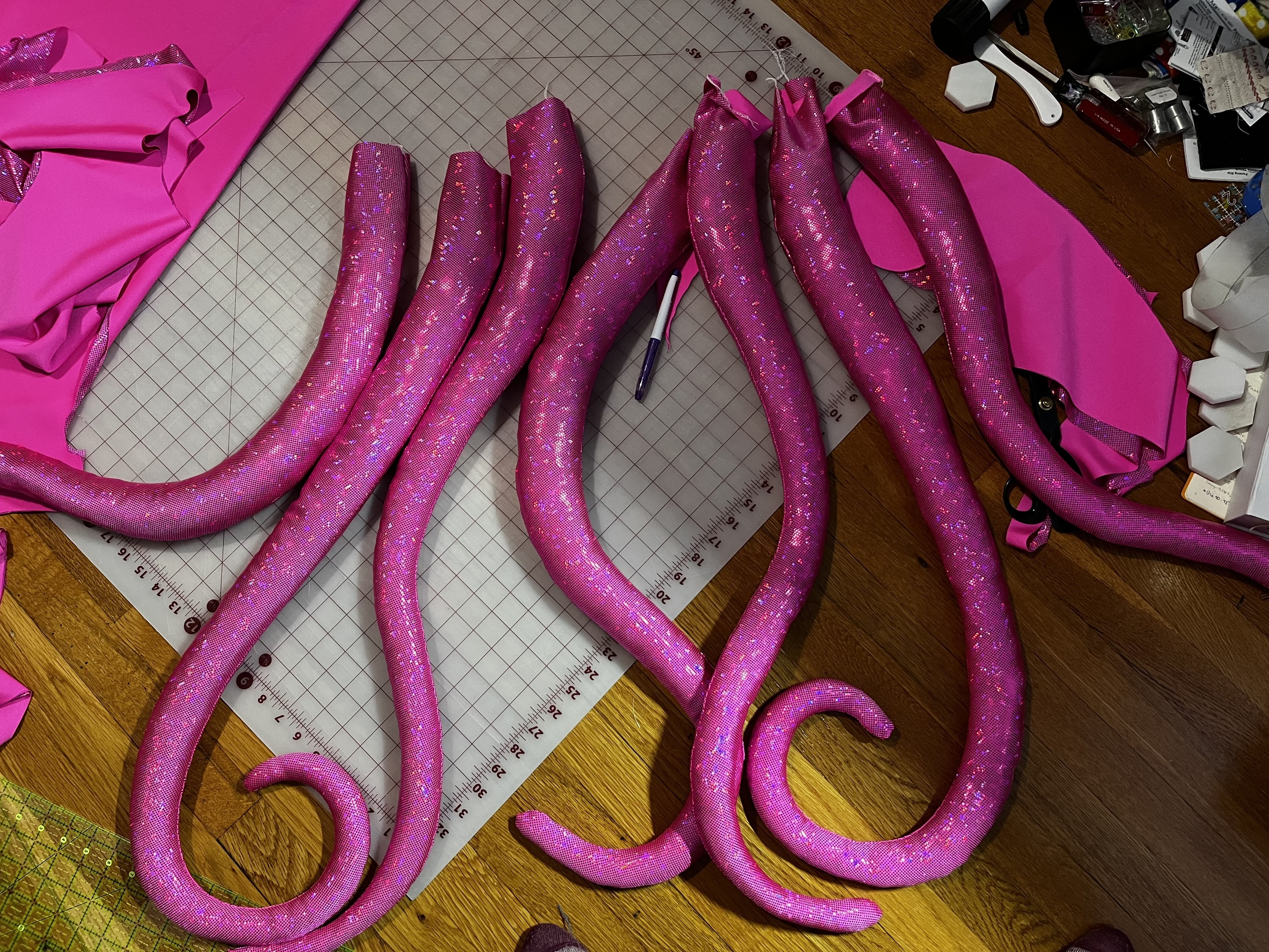Seven hot pink sparkly stuffed tentacles lie on a cutting mat on the floor. They are very  tentacly and glisten in an oozy kind of way.  God I love them.