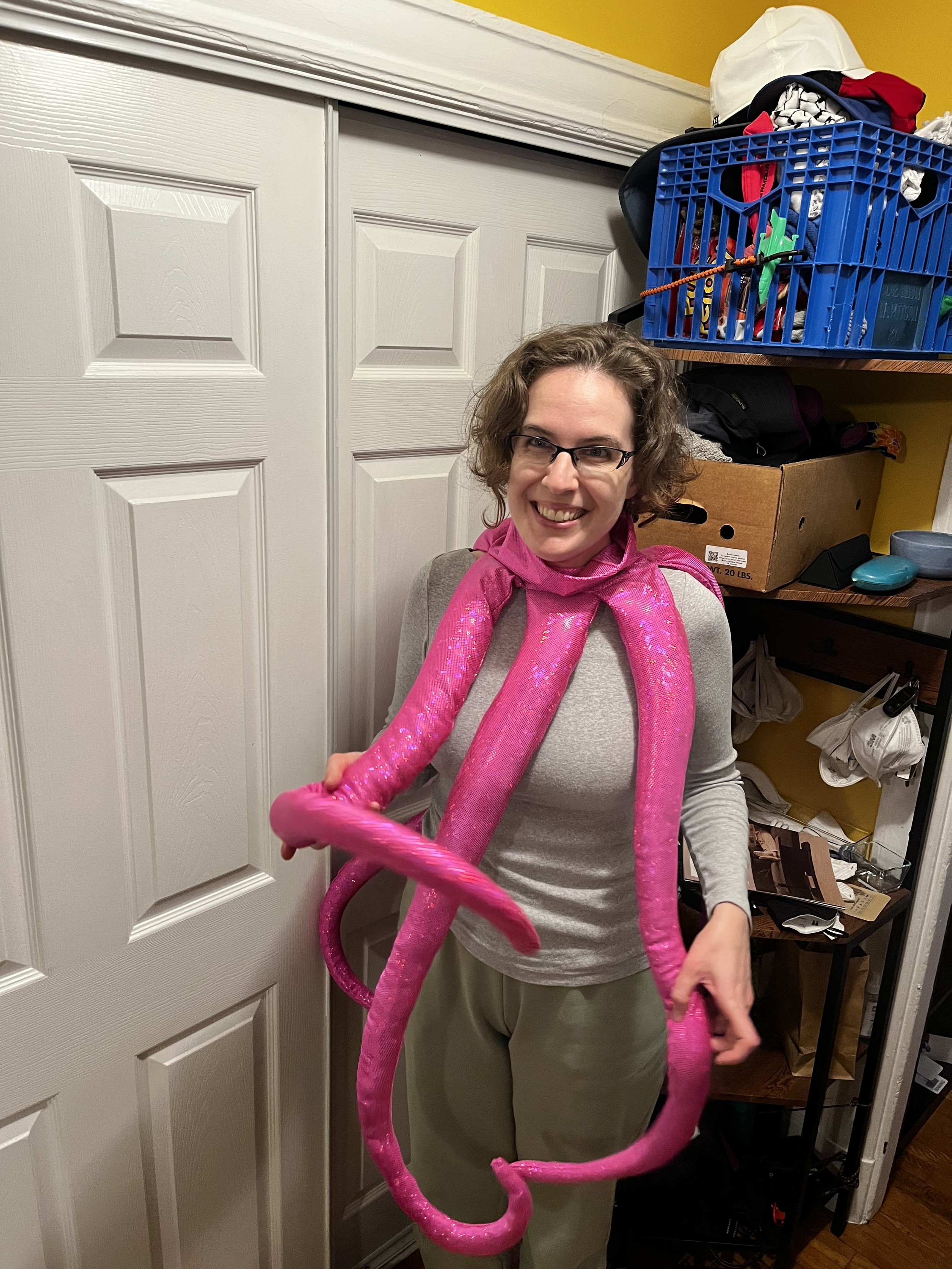 A Caucasian woman is standing in a foyer wearing a hot pink necklace made of thigh-length tentacles. She is grinning madly and the tentacles are all wiggly.