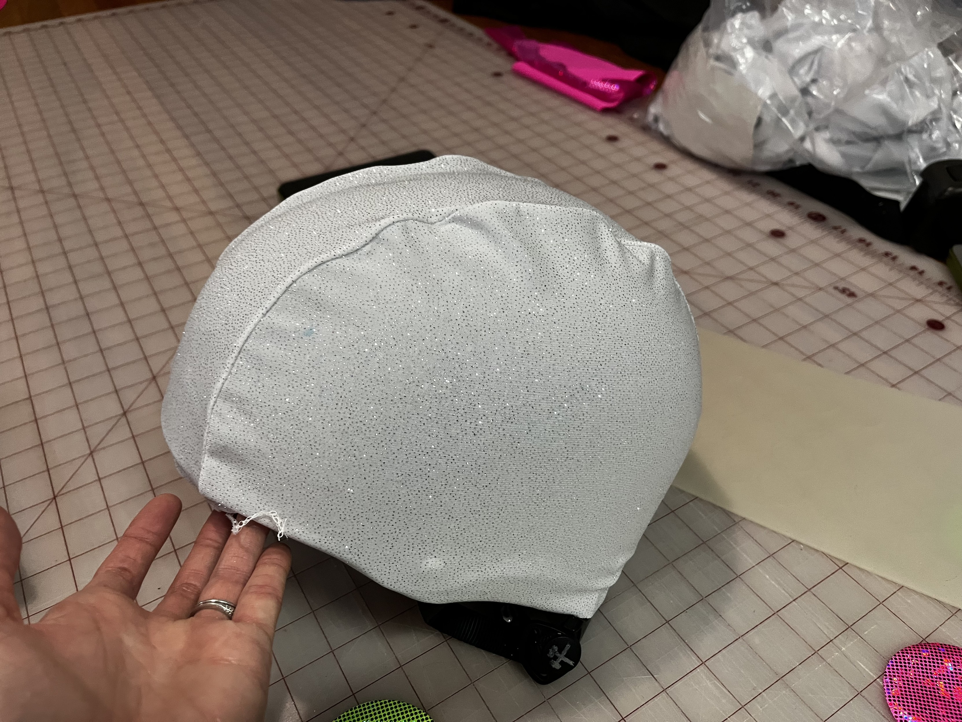 A Caucasian woman’s hand is holding a bike helmet covered with a roller derby style “panty”, a stretchy helmet cover made out of white fabric with very small silver glitter on it. It glistens horribly.