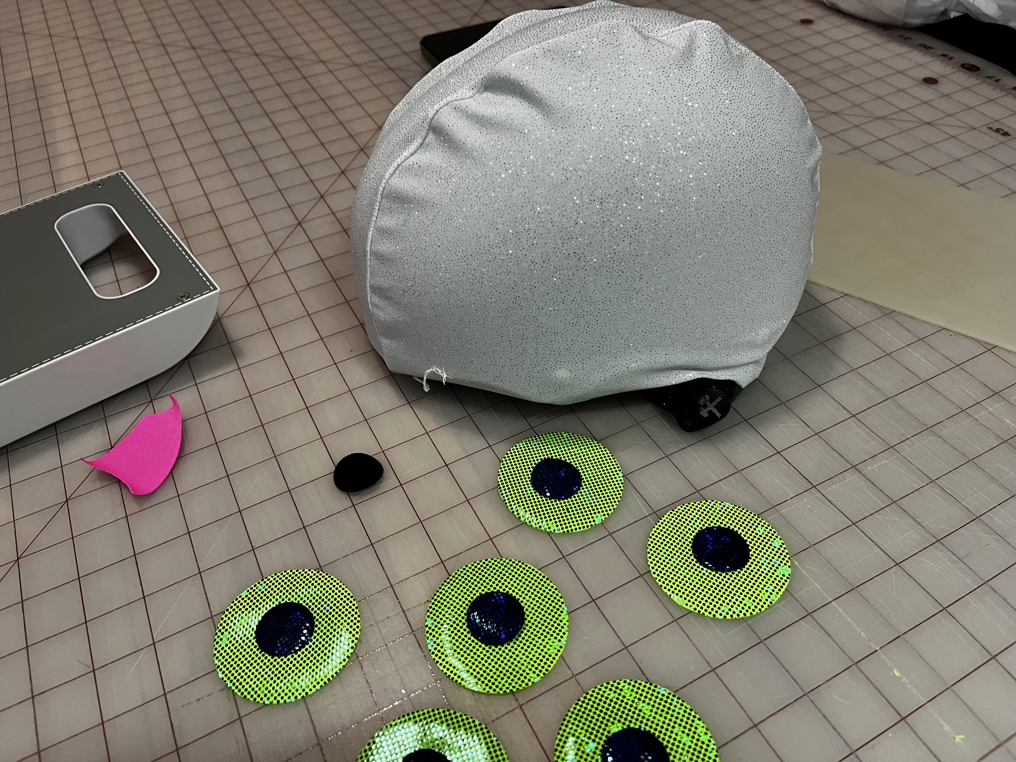The same helmet is on a gridded cutting mat on the floor. In front of it are several of 2.5-inch circles cut out of bright green sparkly fabric with a small blue-black pupil in each one.