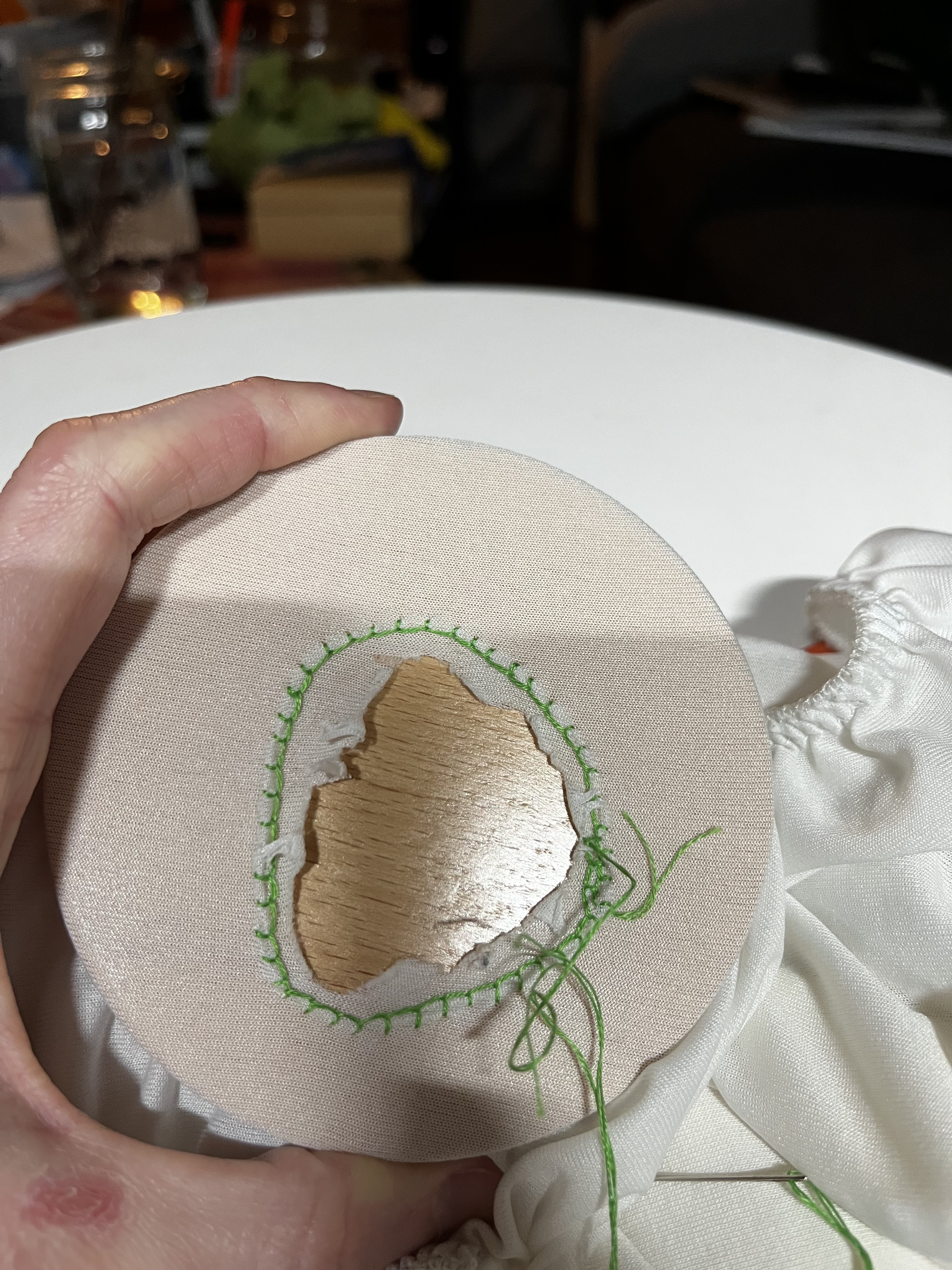 A stretchy white fitted sheet held over a darning surface by a white woman’s hand.  There’s a large jagged hole in the sheet and a rough circle of teal blanket stitch around the perimeter.