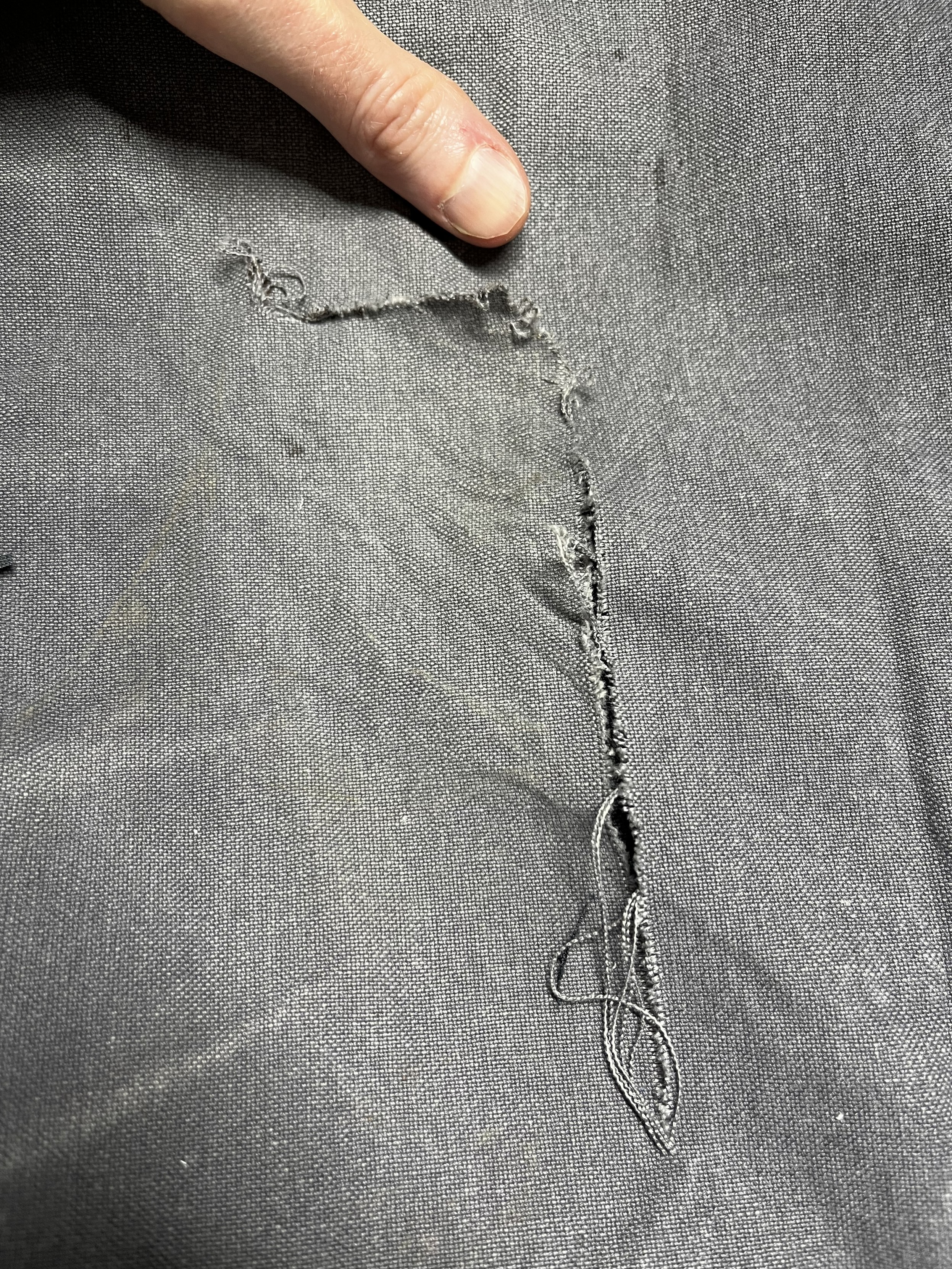 A pair of black (well, formerly black) canvas work pants with a large inverted L-shaped rip in it. A woman’s hand is hiding a bloodstain at the top of the rip.