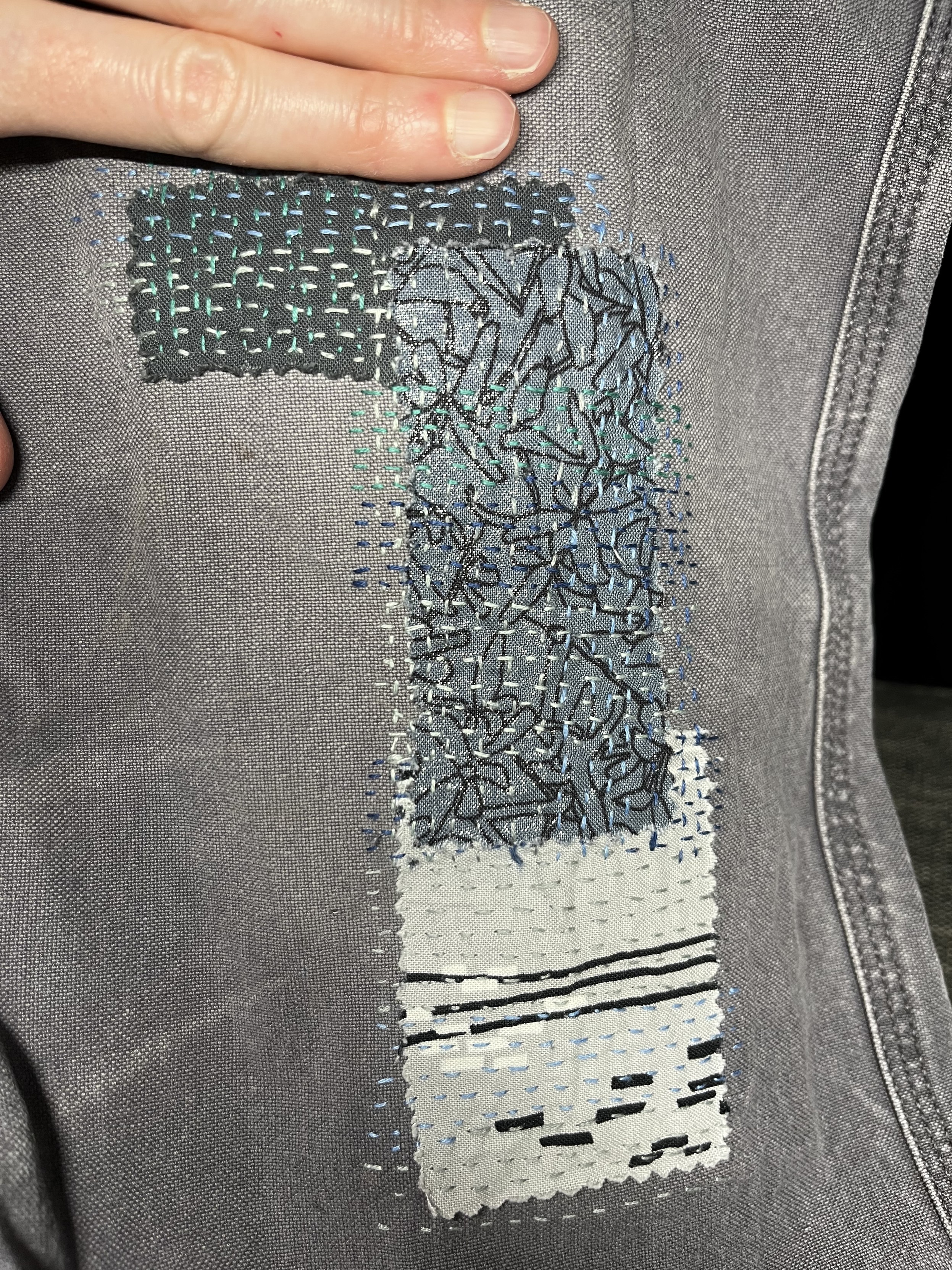 The rip has been mended with three patches in dark grey, silvery black scribbles fabric, and a lighter grey with some black and white lines and random brickwork type shapes on it. Large ish quilting style backstitches cover all three patches in various shades of blue-silver-green.