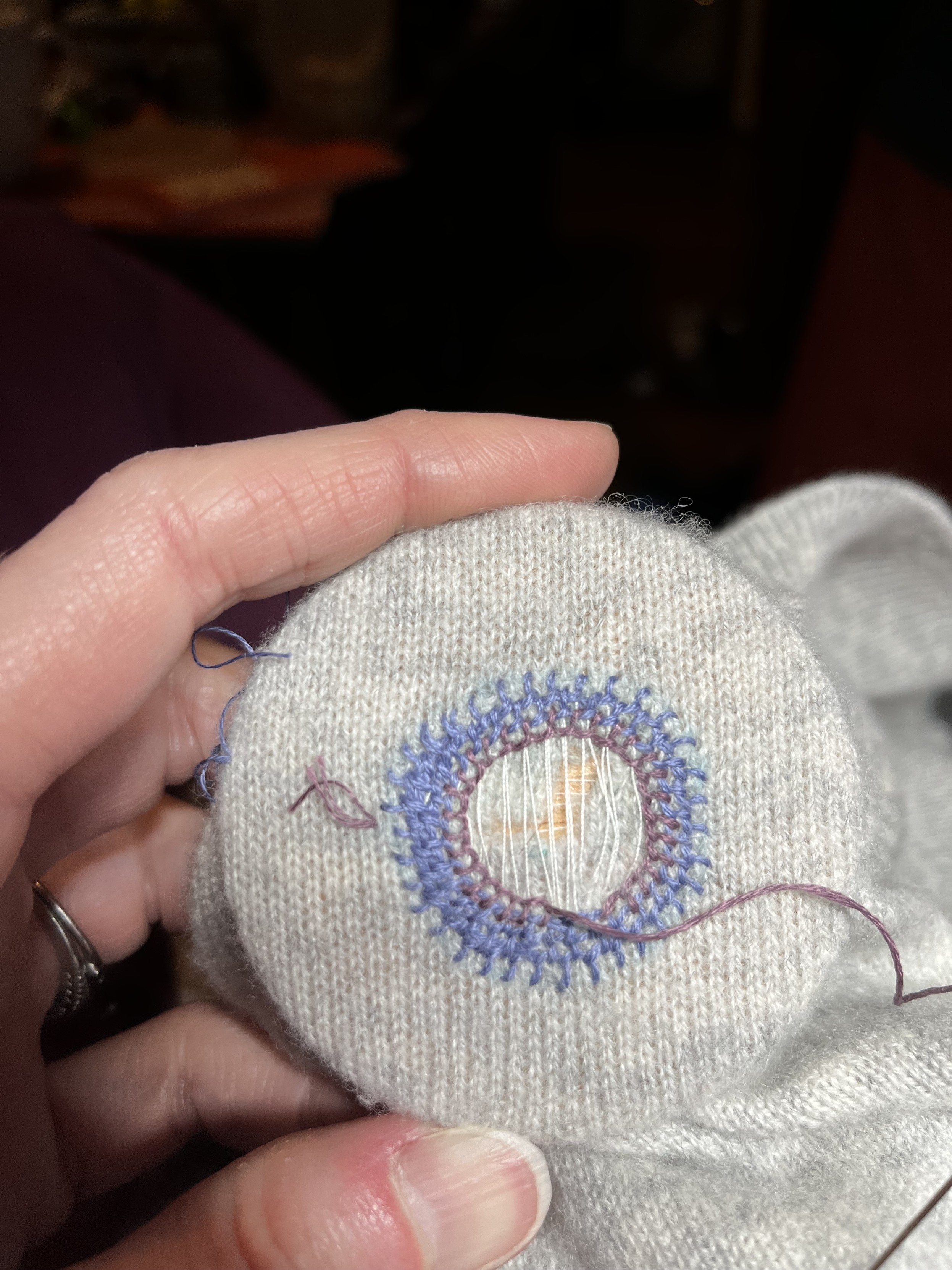 A white woman’s hand is holding a wooden darning surface covered by a light grey sweater in a very fine merino knit. Centered on the darning surface is an l-shaped rip in the sweater. It’s been stitched over loosely with white thread to stabilize it in a cross hatch type pattern, and it’s surrounded by the start of a honeycomb darn (concentric circles of blanket stitch) in a soft blue and then a soft purple.