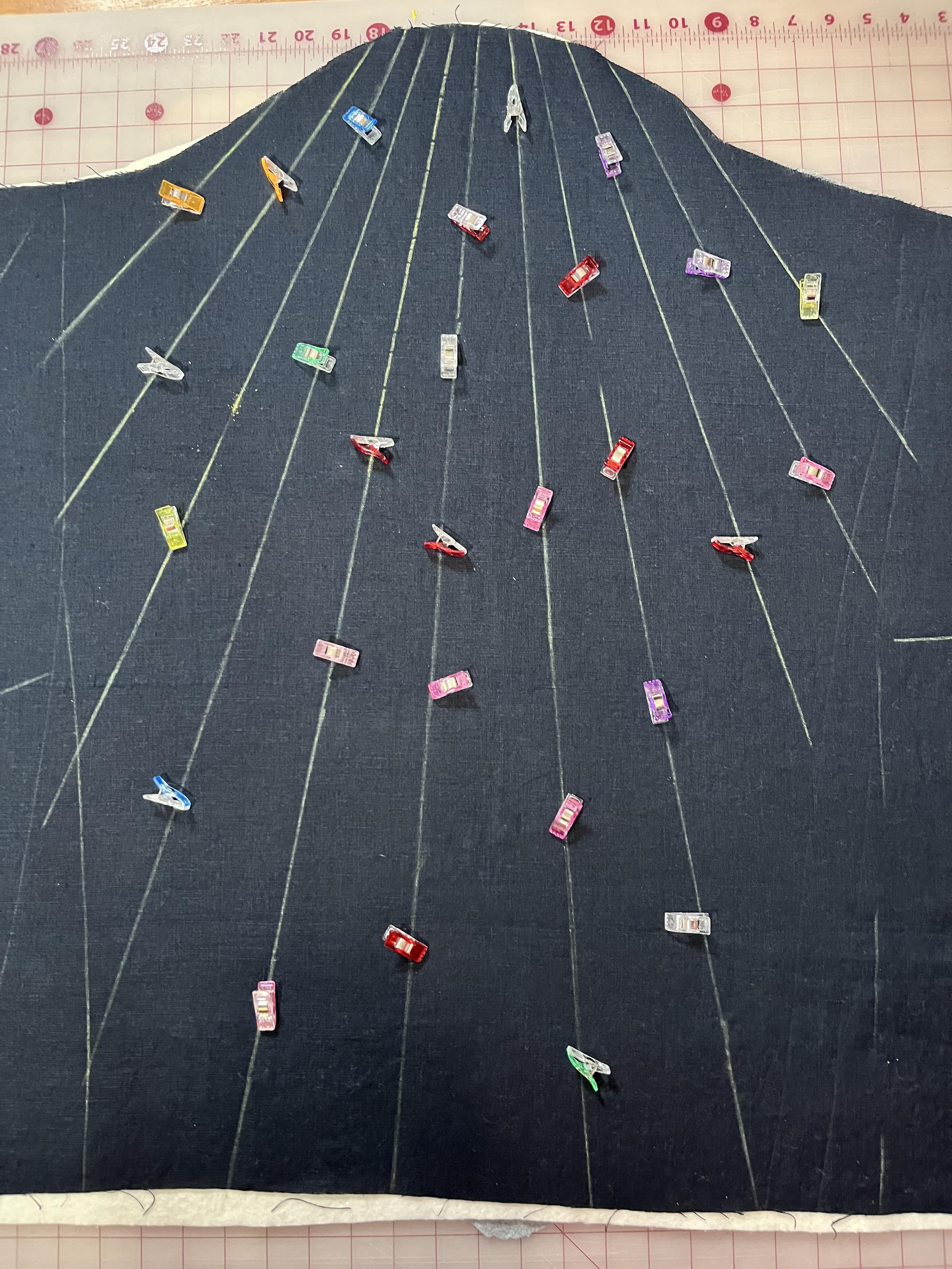 A piece of navy blue linen is on top of a quilt sandwich. The top edge is a vaguely shoulder-cap shaped curve. Radial lines have been drawn in yellow chalk from the head of the shoulder cap and multicolored sewing clips sprinkled on the top and then placed along the radial lines.