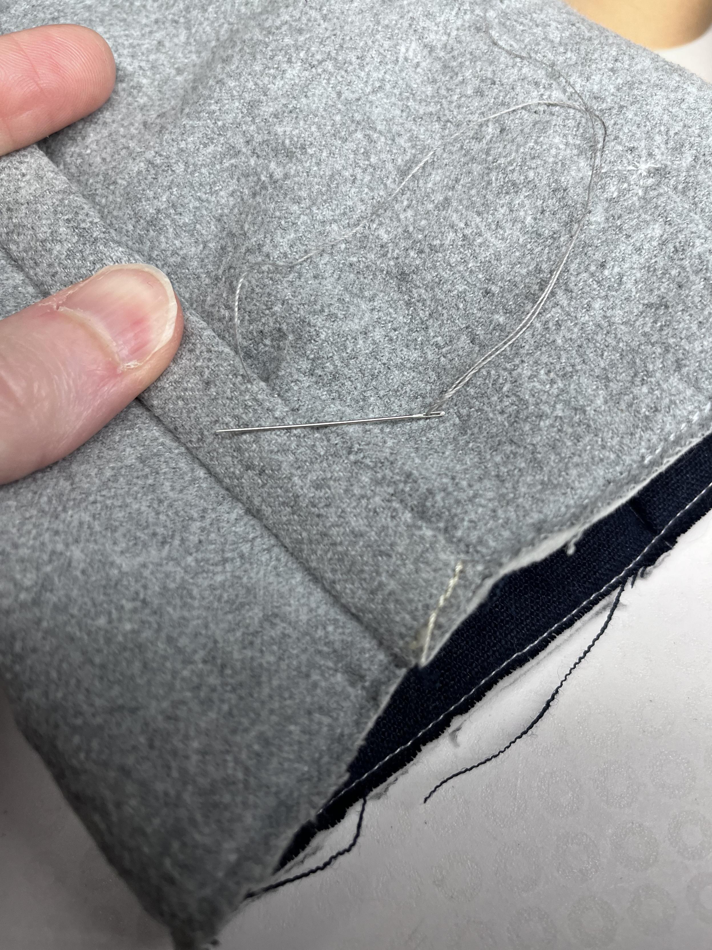 The seam is being held in its final orientation with the binding covering the trimmed seam allowances. It’s partially sewn down with a hand needle visible just below where the seam is being held down.