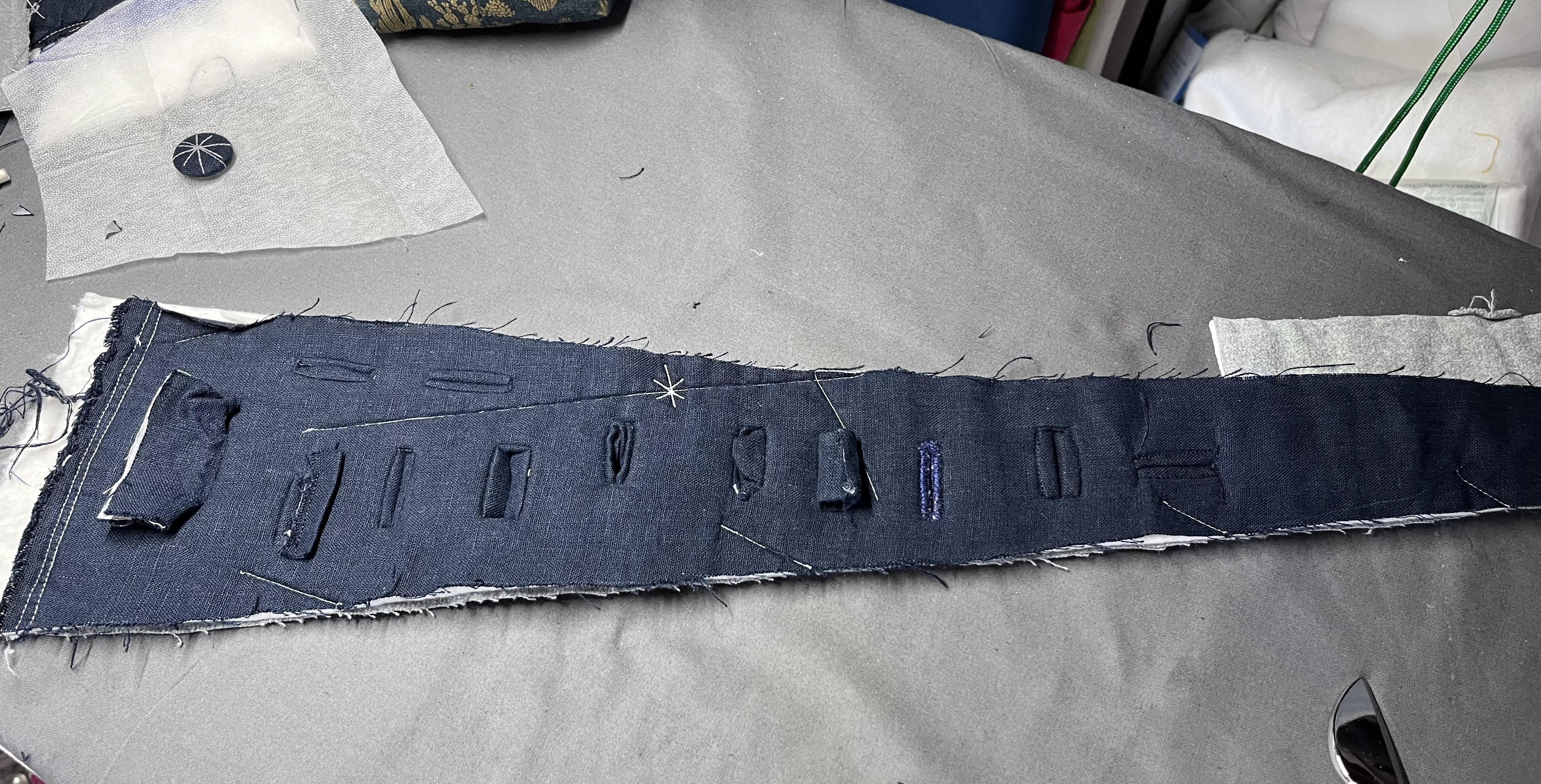 A triangular scrap of quilted fabric with a navy blue linen upper and a grey flannel backing. Twelve different buttonholes have been randomly scattered around it in various states of goodness and doneness. Most of them are very, very bad.